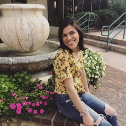 Image Description: Angela is sitting down near a water fountain and smiling at the camera. There are pink and white flowers around her. She is a white woman with shoulder-length dark brown hair, wearing a yellow blouse with flowers, ripped blue jeans, and a pair of black sunglasses on top of her head.