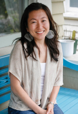 Mia sits on a bench on a front porch. She is Asian American with long wavy hair and smiles for the camera. She is wearing a draped knit sweater.