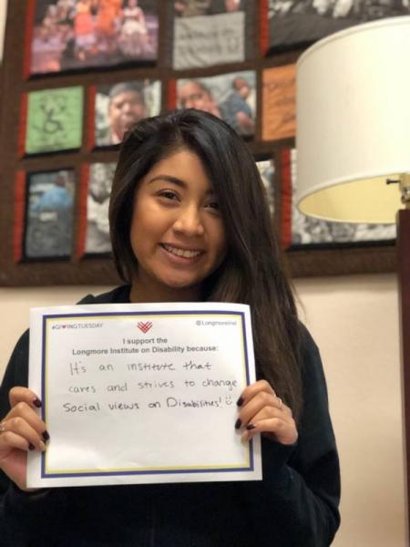 A young woman who appears to be Latina, has long straight brown hair, lightly tan skin, and a big smile, is holding her UnSelfie which says "I support the Longmore Institute because it's an insitution that cares and strives to change social views on Disabilities :)