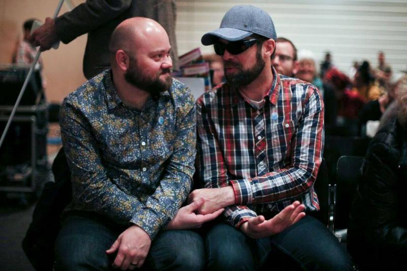 Two white, bearded men at Superfest who are seated closely to each other holding hands. The man on the left gazes at the other affectionately and the man on the right is turned towards him and wears dark glasses.
