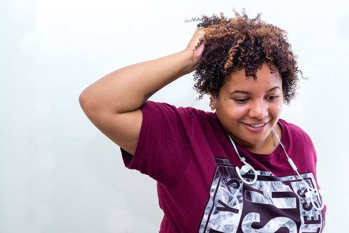 A still of Dani, from "Who am I to Stop it" film who has medium-brown skin, curly dark-brown hair, with ear buds around her neck and a maroon t-shirt.