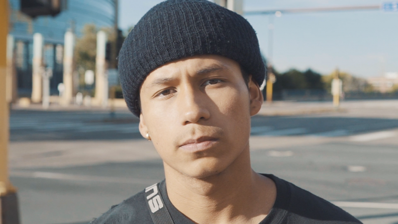 A teenage boy looks into the camera. He has brown skin and is wearing a black beanie and t-shirt.