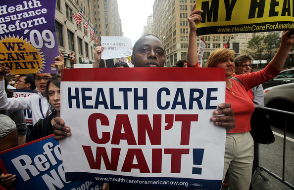 A black man holds a sign at a protest that reads "Health Care CAN'T WAIT!" at a protest