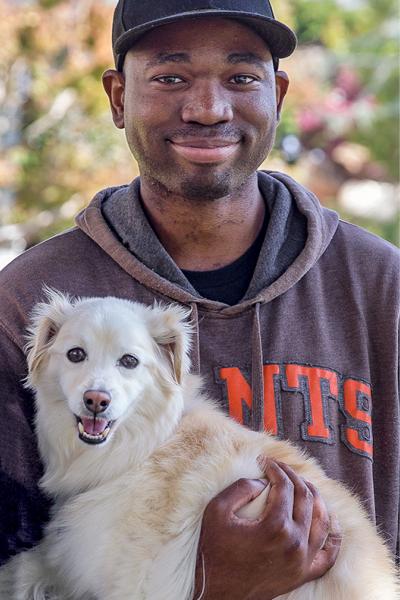 A photo of Lewis who is a black cis man, holding a soft small dog, smiling.