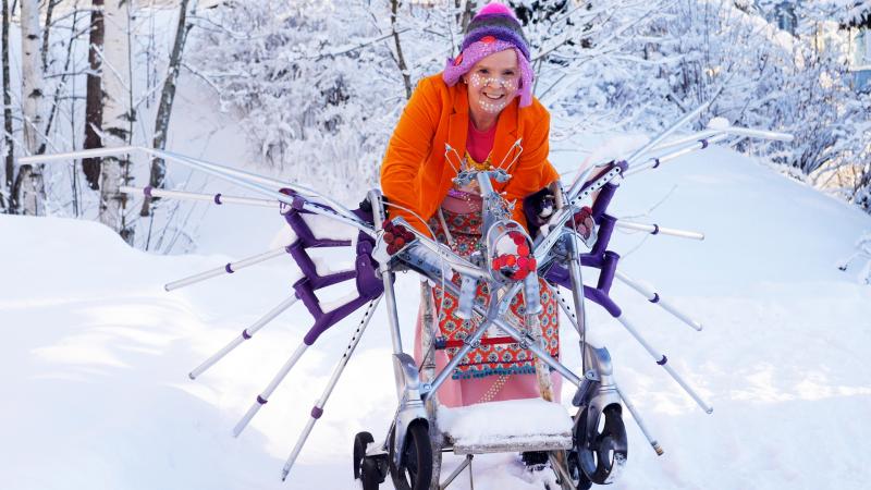 A white woman stands in the snow behind a sculpture made of mobility devices. She is wearing bright colors and white spots on her face. She is smiling.
