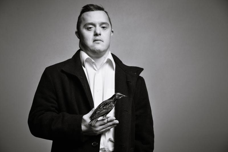 A light skinned man with Down Syndrome is wearing a white button up shirt and dark jacket. He is holding a bird. The whole image is in black and white.