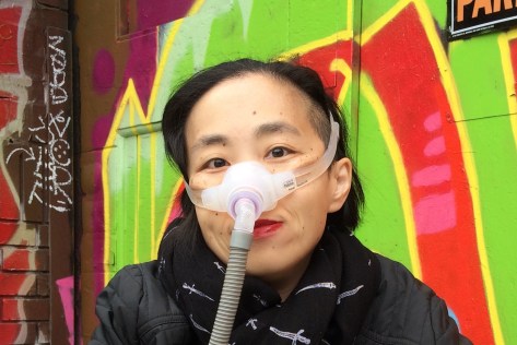 Asian American woman in a wheelchair. She is wearing a black jacket with a black patterned scarf. She is wearing a mask over her nose with a tube for her Bi-Pap machine. Behind her is a wall full of colorful street art