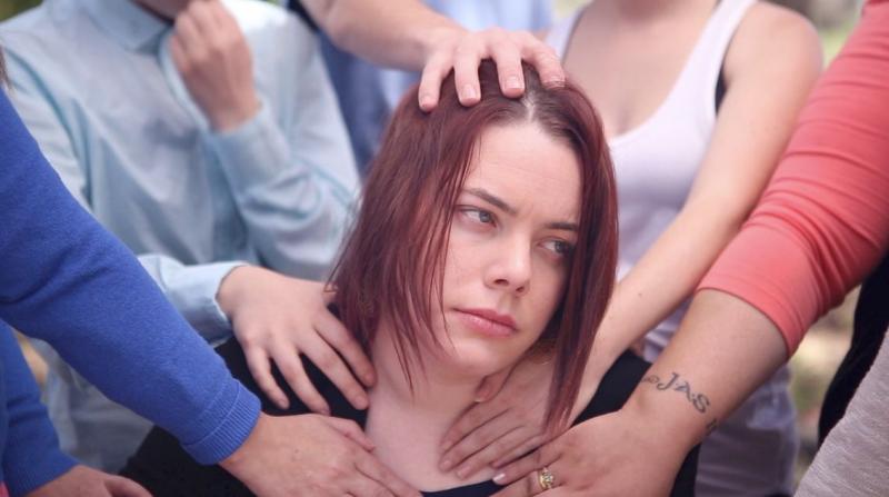A white woman with red hair looks annoyed. There are many hands touching her head and shoulders.