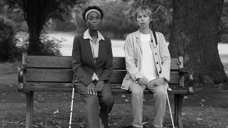 Two women sitting on a bench, one woman is Black and one is white. They both have canes and are looking off into the distance.