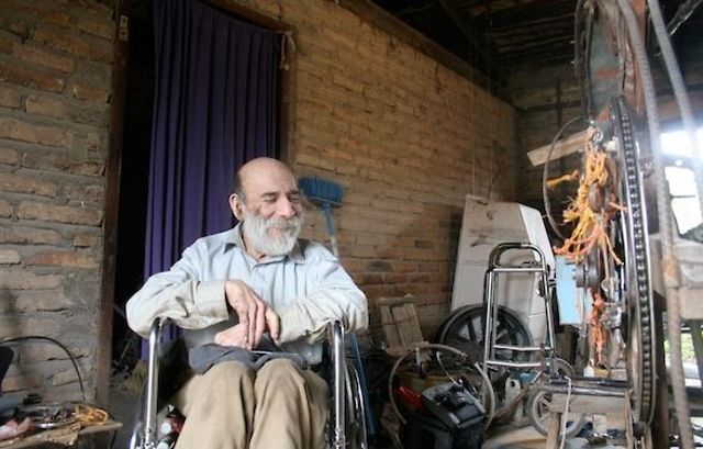 An elderly man in a wheel chair closes his eyes and smiles at the camera as he is surrounded by bike parts, scrap metal, and old tires.
