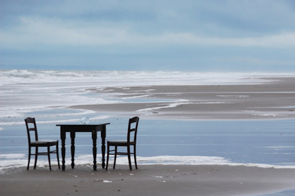 The silhouette of a table sits between two chairs on a beach during low tid