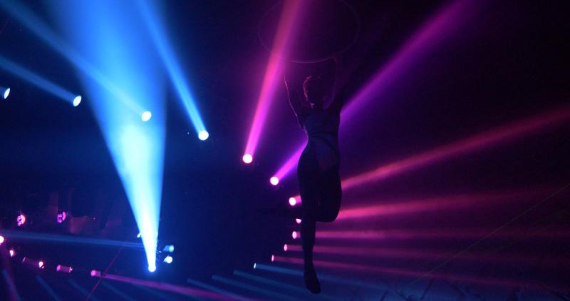 A person hanging from a hoop. Their body is silhouetted by bright pink and blue lights.
