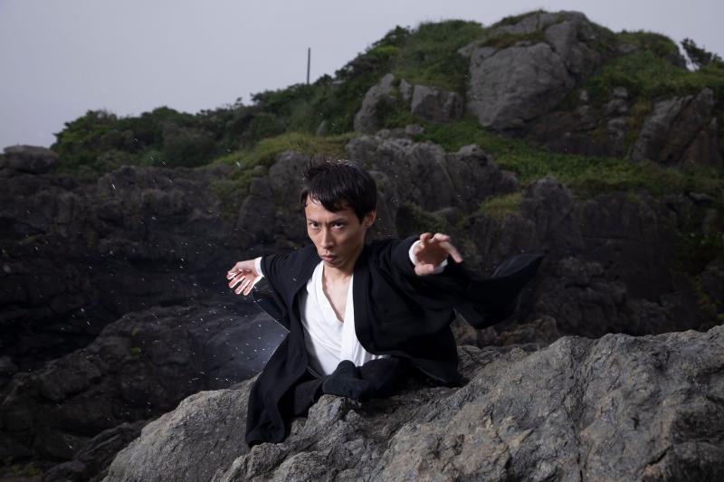 A Japanese man sits on a rock formation and is gesturing toward the camera. He is wearing black and white flowy clothing.