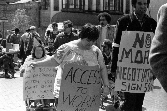 People protesting before entering the building. A prominent sign a woman is wearing around her neck reads "Access to Work"