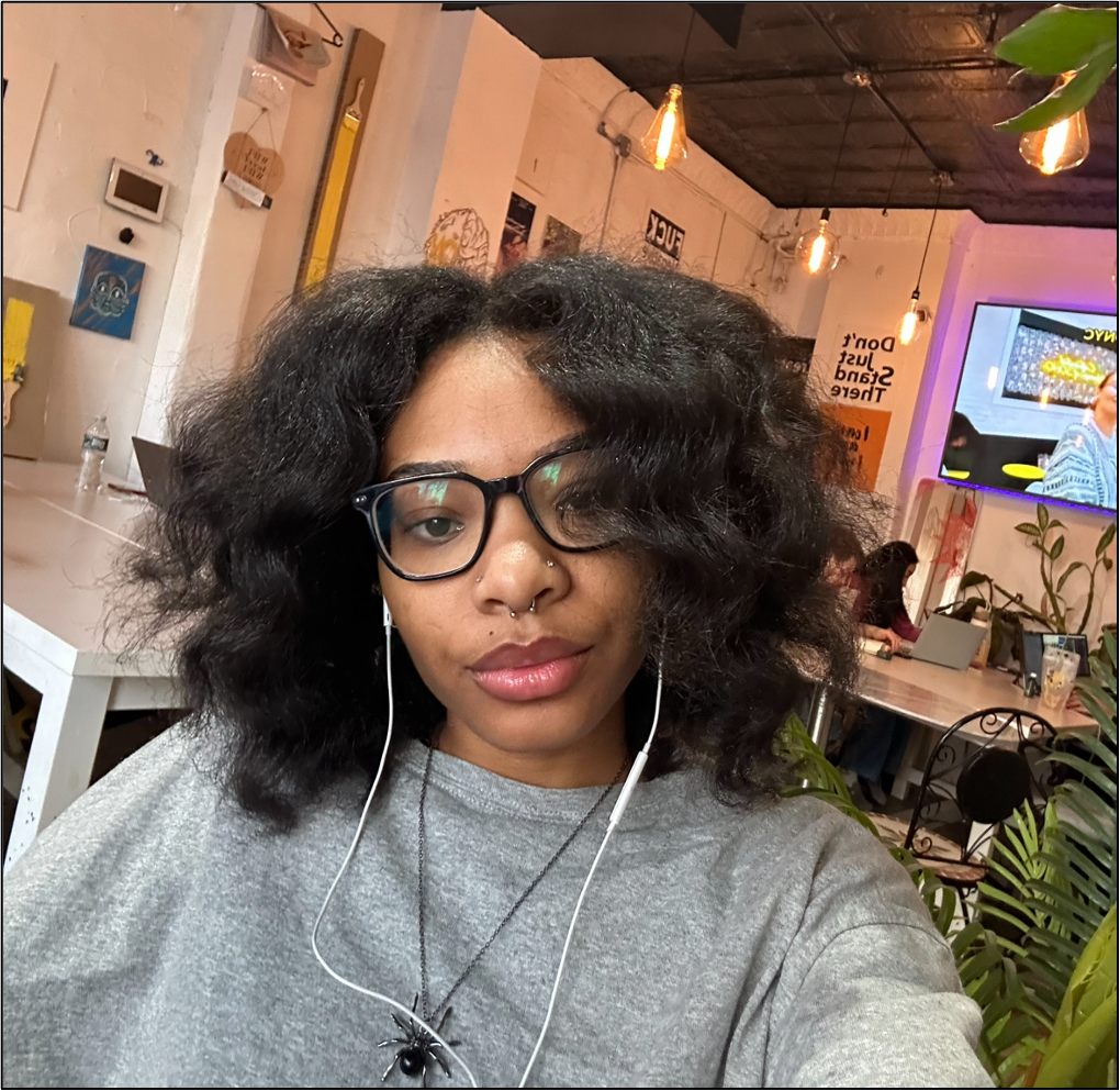 A young Black person with curly black hair takes a selfie in a warmly lit café. They wear black-framed glasses, eyebrow and nose piercings, a gray oversized shirt, and a black spider necklace, with white earbuds in their ears. The background features hanging lights, wall art, a television screen, and people working on laptops.