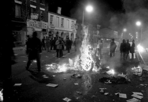 a wheelchair burning up in flames, surrounded by a crowd