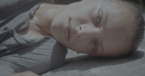 A young woman with light skin and light eyes wearing a grey shirt lies on the ground on her side, looking at the camera.