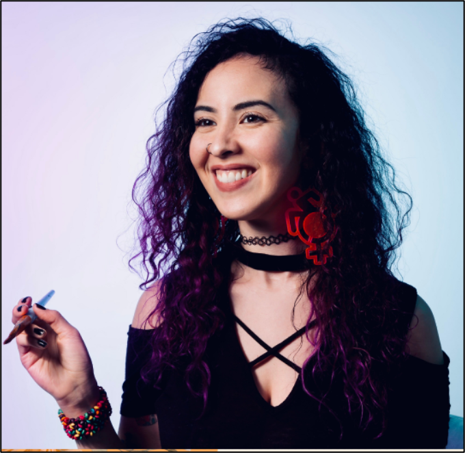 Genevieve, a light-skinned Mexican woman smiles before a bold periwinkle background, holding up a paintbrush in one hand. Her black curly hair is long, past her shoulders. She wears a black dress and black choker necklace.