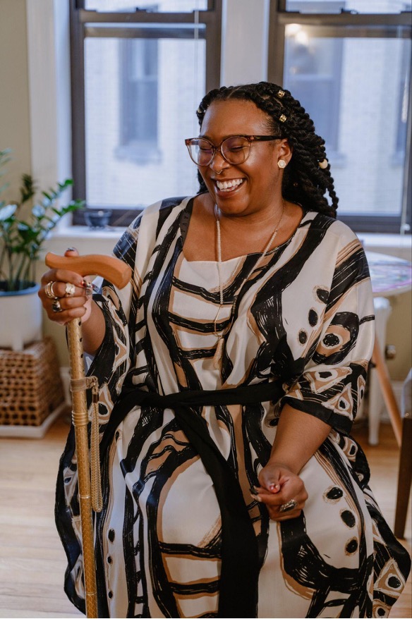 Jade T. Perry, a BlackQueerDisabled femme, sitting with their wooden cane and laughing joyously with their eyes closed and mouth slightly open. She wears a black, brown, and cream flowing dress and brown glasses.