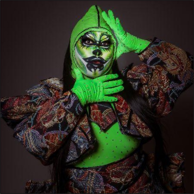 OBSIDIENNE OBSURD, a Chinese-American drag queen, stands in a fierce model-esque pose, with a white makeup base, face painted in greens, yellows, articulated with black linework. they are wearing a tapestry horse-print crop suit jacket and matching skirt, with a like green stoned full bodysuit underneath. They stand posed wearing a green hood with a slick, shiny ponytail, split to rest on the shoulders.