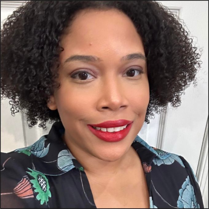 Tiezst “Tie” Taylor, a light skinned Black femme, appears in the photo wearing a dark blue collared dress with blue, green, and orange floral accents. They have dark Brown curly hair and are wearing dark red lipstick and pink eye shadow. They are standing against a white background.