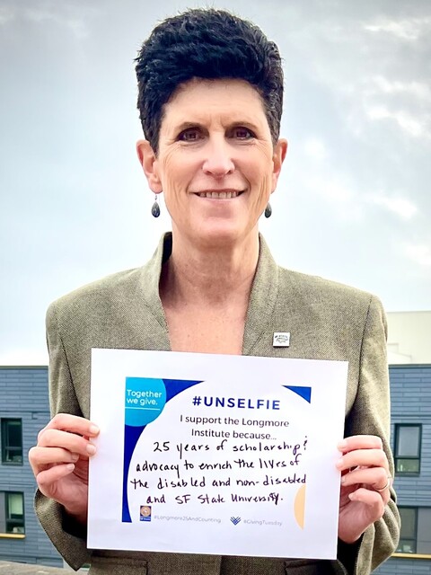 San Francisco State University President Lynn Mahoney. She is a white woman with short dark hair. She holds a sign that reads, “I support the Longmore Institute because of 25 years of scholarship and advocacy to enrich the lives of the disabled and nondisabled and SF State University.”