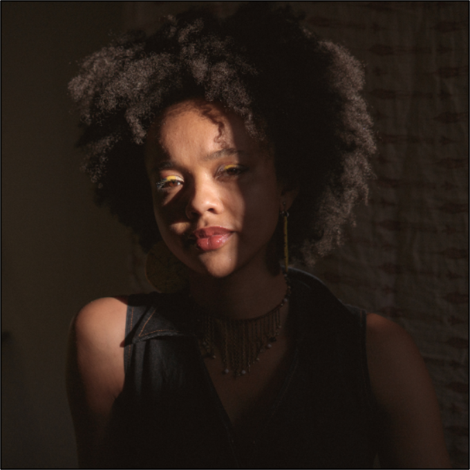 peace, a light skinned Black person, gently looks at the camera, their face partially in shadow. Their hair is a medium afro a curl creates a shadow on their face. They wear large yellow beaded earrings and a brown beaded necklace.