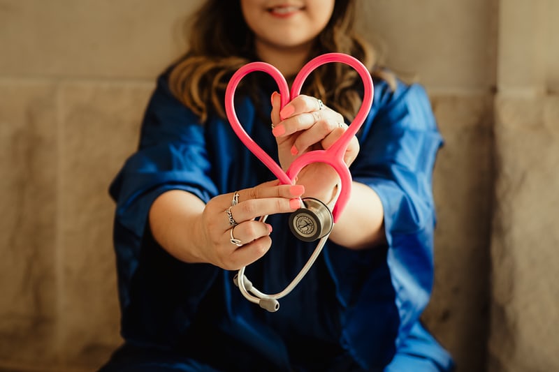 A person with long hair in blue scrubs shapes the tubing of a stethoscope into a heart