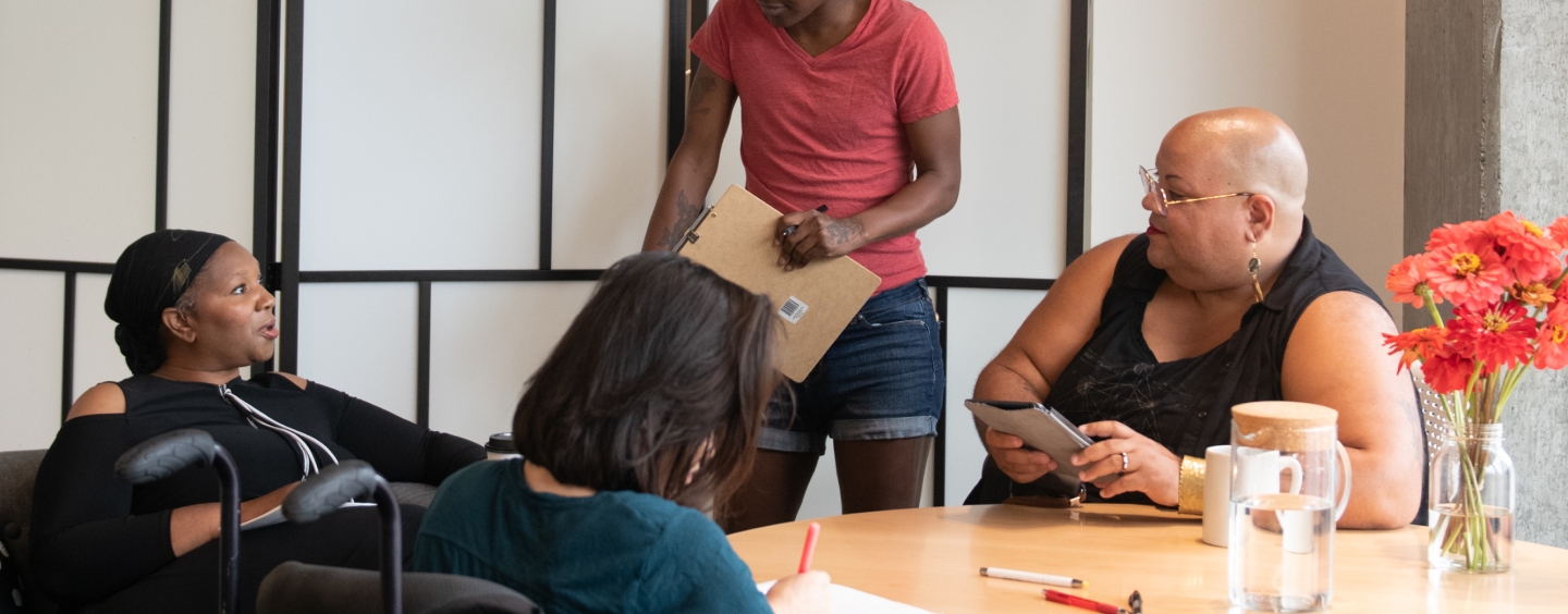 Four disabled people of color gather around a table during a meeting. A Black woman sitting on a couch speaks with a neutral expression while the three others (a South Asian person sitting in a wheelchair and taking notes, a Black non-binary person sitting in a chair with a tablet and cane, and a Black non-binary person standing with a clipboard) listen.