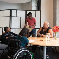 Four disabled people of color gather around a table during a meeting. A Black woman sitting on a couch speaks with a neutral expression while the three others (a South Asian person sitting in a wheelchair and taking notes, a Black non-binary person sitting in a chair with a tablet and cane, and a Black non-binary person standing with a clipboard) listen.