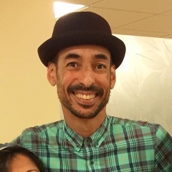 Robert, a person with light brown skin and facial hair, smiles at the camera in a zoomed-in shot. He wears a black hat and a green plaid button-up shirt. 