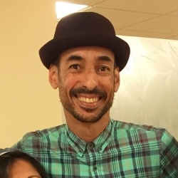 Robert, a person with light brown skin and facial hair, smiles at the camera in a zoomed-in shot. He wears a black hat and a green plaid button-up shirt.