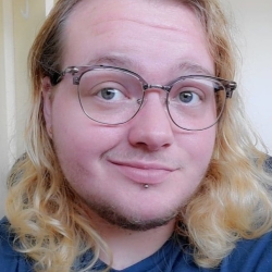 Oleander, a person with light skin, smiles at the camera wearing glasses, a blue shirt and has blonde wavy hair. 