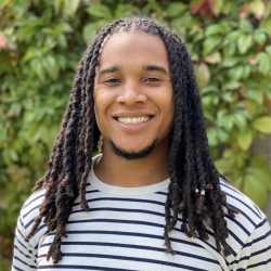 A close portrait of Philip, a person with dark skin and long hair, smiling in front of green foliage wearing a black and white striped shirt.