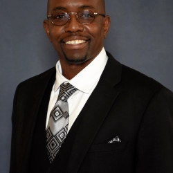 Omar, a person with dark skin and facial hair, smiles at the camera and stands in front of a grey backdrop. He wears glasses, a grey tie, and a black suit jacket. 