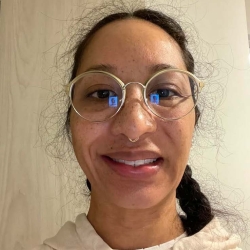 A close portrait of Bernadette smiling. They wear a cream-colored hoody, wireframe glasses and their dark brown hair is braided. Bernadette has light brown skin and a septum piercing.
