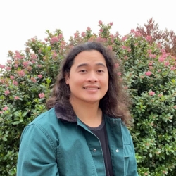 Nam smiles in front of a green bush with red flowers. He has long wavy brown hair and wears a teal jacket. 