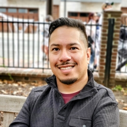 Headshot of Matthew Alaniz, a Latino man with black slicked back hair and short facial hair. He is outside in front of a fence, wearing a grey jacket.