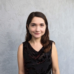 Mia, light-skinned and with brown hair, stands in front of a grey wall wearing a black shirt with a velvet design. She smiles at the camera.