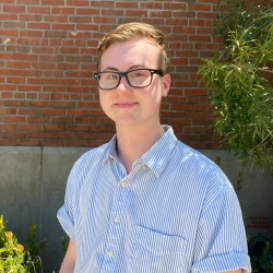 Nathan Burns stands in front of a brick wall, smiling for the camera. They are a white person with short blond hair, and are wearing glasses and a blue and white striped shirt.