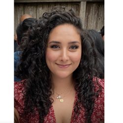 Iyah, a Southwest Asian North African woman, smiles at the camera in a close portrait. Wearing a red floral dress and has dark brown curly hair.