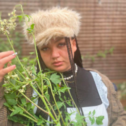 Image Description: Bayley stands close to the camera wearing a fuzzy hat, black turtleneck, and a plaid shirt layered on top. She holds a green plant. 
