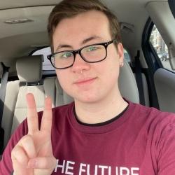 A selfie of Nathan Burns, a white person with short blonde hair and glasses. They are holding up a peace sign, and wear a shirt that says "The Future is Disabled."