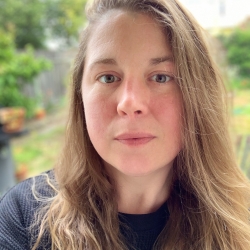 A person with light skin and long brown hair. Katie is wearing a black shirt and standing outside in front of some greenery.