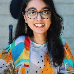 Shaina, with long black hair, dangly gold earrings and round black framed glasses, is a South Asian woman, whose wheelchair headrest shows behind her head. She's wearing a vibrant shirt with swervy colors and black spots.