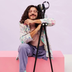 Alex, a Black biracial amputee smiles while propped against their black crutches. They have a pastel floral print blazer, a double string of pearls, purple pants, and shoulder length loose black curls. 