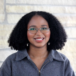 A headshot of a grinning Alana, a Black woman with shoulder length coily hair, glasses, and light red lip gloss. She stands in front of a white stone wall.