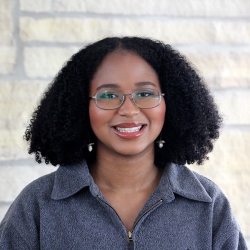 A headshot of a grinning Alana, a Black woman with shoulder length coily hair, glasses, and light red lip gloss. She stands in front of a white stone wall. 