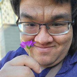 On a sunny day, Chris, wearing eyeglasses and a purple shirt, smiles at the camera and holds up a small purple flower.