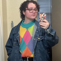 Joe is a round-faced white person with medium length brown curly hair and dark blue glasses. They are standing in a bathroom with beige walls, wearing a denim jacket, jeans, and a rainbow checkered sweater. They are holding their phone up to a mirror and looking into the camera with a neutral expression.